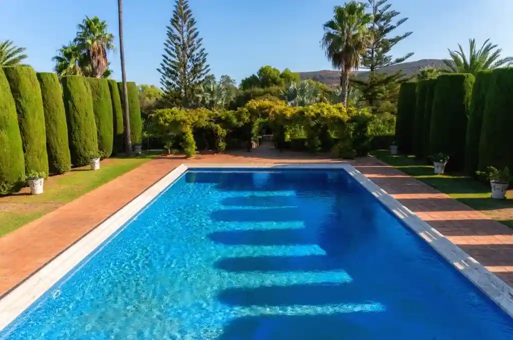 Blick auf ein Schwimmbad mit kristallklarem Wasser, umgeben von Gärten, an einem sonnigen Morgen.